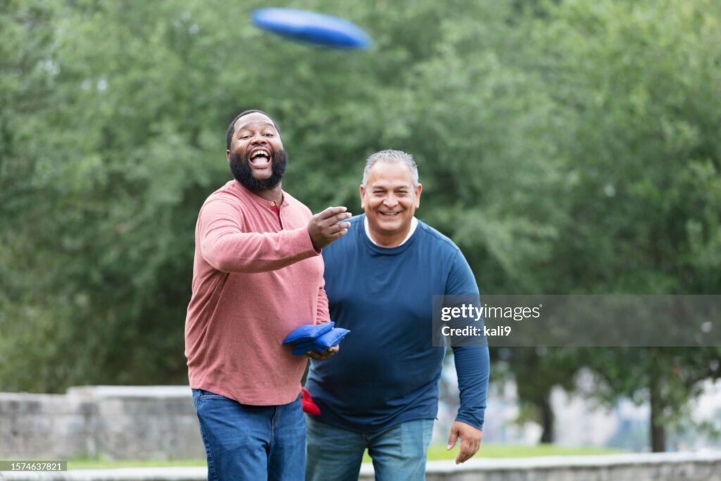 Two plus size multiracial men having fun at the park playing a bean bag toss game. The main focus is on the African-American man, in his 30s, who has tossed his bean bag and is shouting and cheering, waiting for it to land. His Hispanic friend is behind him, watching and smiling.